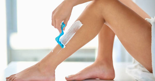 A woman carefully shaves her legs with a razor, contemplating whether to exfoliate before or after shaving.
